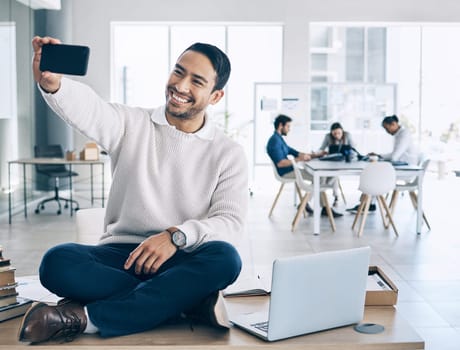 Selfie, phone and businessman sitting on desk in meeting with team, staff and working in corporate office. Startup, happiness and young Asian male worker using smartphone to take picture in workplace.