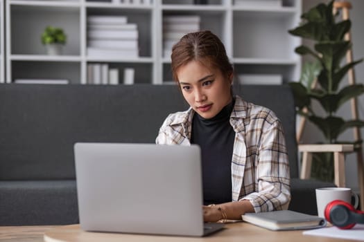 Beautiful and inspired Asian woman working on her tasks on laptop in a living room on the weekend...