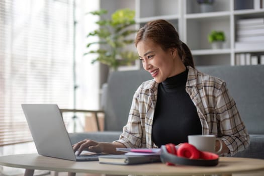 Beautiful and inspired Asian woman working on her tasks on laptop in a living room on the weekend...