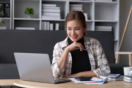 Beautiful and inspired Asian woman working on her tasks on laptop in a living room on the weekend...