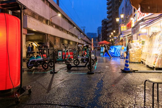 Osaka, Japan - April 29, 2023: Electric scooters and bikes parked by bright drug store and train station on rainy night. High quality photo
