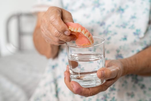 Asian senior woman patient holding and washing denture in water cleanser glass for good chewing.