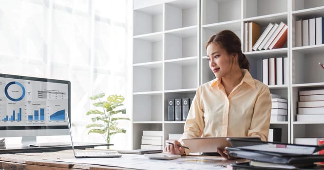 Beautiful and happy young Asian businesswoman looking at her laptop screen while enjoying her morning coffee at her desk in the office...