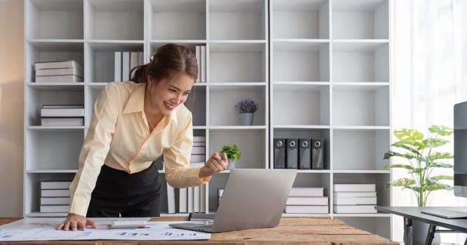 Excited business asian woman sitting at table with laptop and celebrating success..