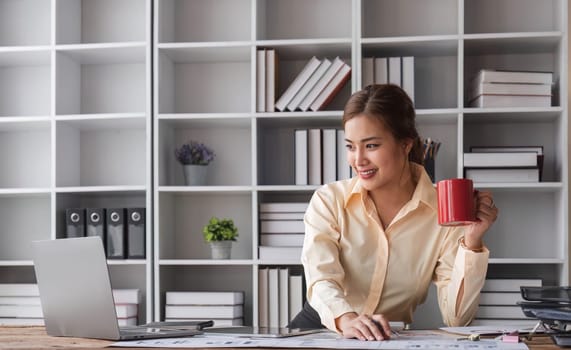 Beautiful and happy young Asian businesswoman looking at her laptop screen while enjoying her morning coffee at her desk in the office...