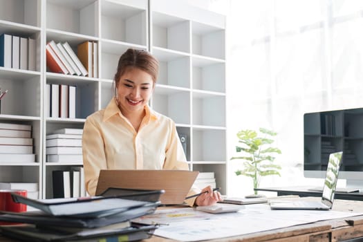 Beautiful and happy young Asian businesswoman looking at her laptop screen while enjoying her morning coffee at her desk in the office...