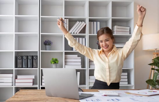 Excited business asian woman sitting at table with laptop and celebrating success..