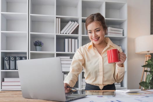 Beautiful and happy young Asian businesswoman looking at her laptop screen while enjoying her morning coffee at her desk in the office...