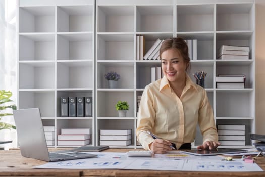 Beautiful and happy young Asian businesswoman looking at her laptop screen while enjoying her morning coffee at her desk in the office...