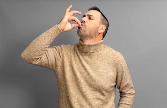 Bearded Hispanic man in his 40s wearing a beige turtleneck smacking his lips and licking his fingers after devouring a delicious treat, isolated on a gray studio background