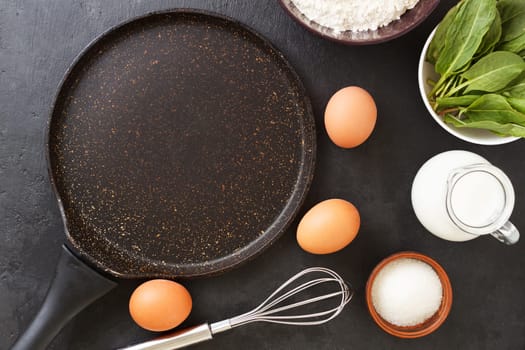 An empty pan for making spinach pancakes on a black background with ingredients: eggs, spinach, milk, flour.