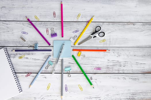 Bright, multicolored pencils for drawing in an album, colored paper clips, a blue notepad for notes, scissors, a pen, a metal sharpener and clips are folded neatly in a circle on wooden background.