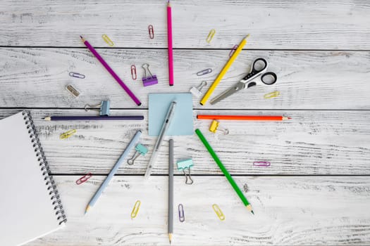 Bright, multicolored pencils for drawing in an album, colored paper clips, a blue notepad for notes, scissors, a pen, a metal sharpener and clips are folded neatly in a circle on wooden background.