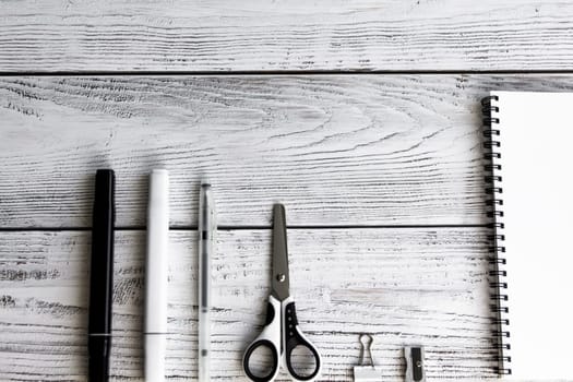 A sketchbook with white sheets, a clip, a metal sharpener, a black and white marker, a writing pen and scissors in black and white tones and shades on wooden background.