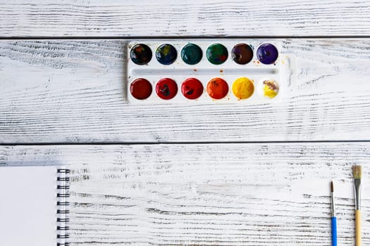 A sketchbook with white sheets, paint brushes and colorful, bright watercolor paints on a light, gray and wooden background.