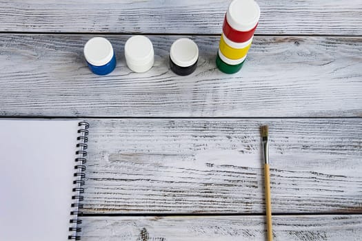 A sketchbook with white sheets, paint brushes and colorful, bright gouache paints on a light, gray and wooden background.