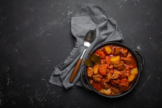 Beef meat stew with potatoes, carrot and delicious gravy in black casserole pot with bay leaves with spoon on black dark rustic concrete background from above, space for text.