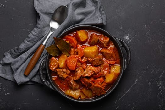 Beef meat stew with potatoes, carrot and delicious gravy in black casserole pot with bay leaves with spoon on black dark rustic concrete background from above .