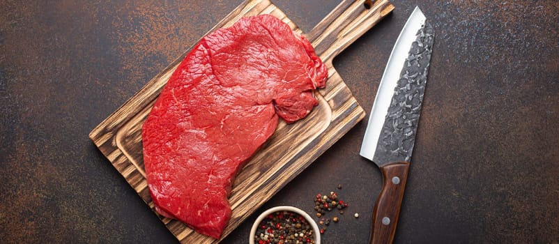 Raw uncooked top round beef steak on wooden cutting board with big kitchen knife and pepper on dark brown rustic stone background top view, cooking meat steak concept