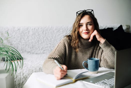 Stylish and focused female PR manager creating content plan for online store, working from home with laptop, notebook and coffee, looking at camera.