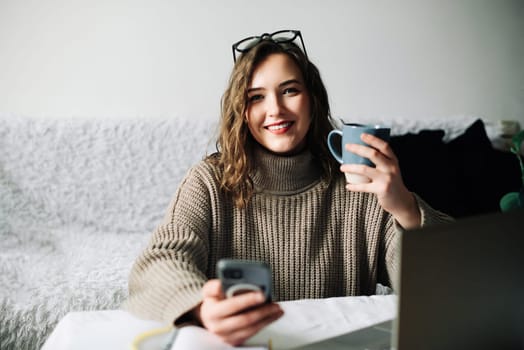 Young student taking a break from online studies, enjoying coffee and phone, laughing at memes and relaxing at kitchen table.