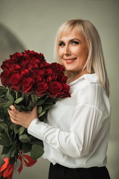 A fashionable woman in a white shirt and black trousers poses in the interior with a bouquet of red roses.