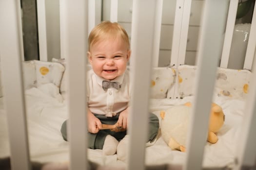 Portrait of a cheerful little boy in a white shirt with a bow tie sitting in a crib.