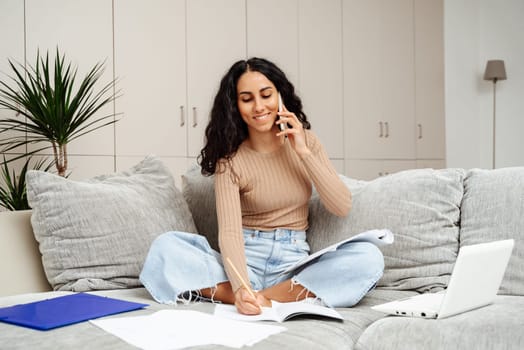 Young beautiful female student of Arab appearance sits on the sofa at home and studies. She holds a phone in one hand and takes notes in the other. Carries on a pleasant conversation about the subject of the task.