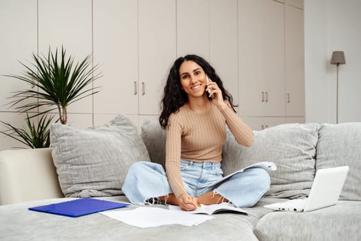 Young beautiful arab girl gets education online. In one hand she holds the phone and calls her teacher with the other hand makes notes. Looks straight ahead smiling brightly.