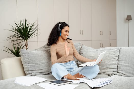 A young beautiful girl of Arab appearance leads a video call. Greeting waving hand and holding a laptop. The girl is sitting on the couch in a homely atmosphere around her a lot of papers.