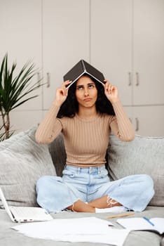 Indoor shot of tired woman wearing casual clothing sitting on sofa and working on notebook, felling fatigue. She covered herself with a notebook and hides like a house from a large amount of paperwork