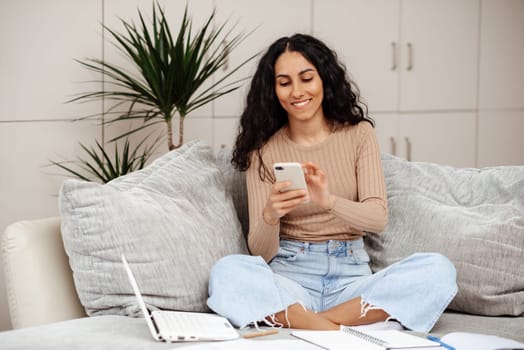 Smiling millennial mixed race woman looks at phone, reads message, has meeting, work with laptop in living room interior. App for communication remotely, study, freelance, good news at home