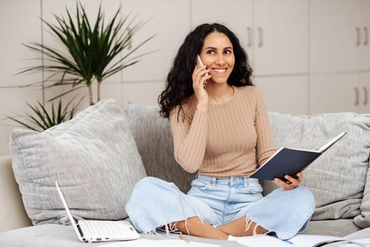 Smiling arabic millennial woman talking on phone holding note book sitting on the sofa in home office. Young beautiful girl makes a work call looks straight ahead around her papers and a laptop.