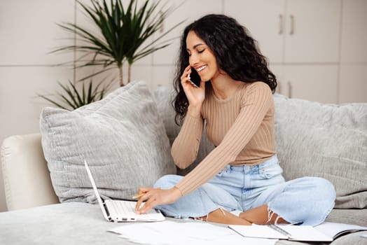Online education concept. Happy middle eastern woman making call about task, studying at home with laptop, taking notes while watching webinar or lesson.