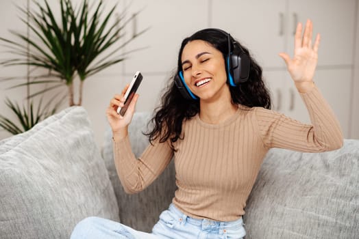 Portrait of a happy young Arab woman listening music with wireless headphones from music application on mobile smart phone in living room at condominium. Teen dancing listening to favorite playlist