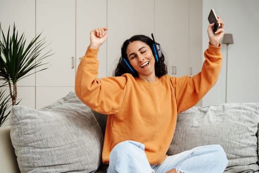 Carefree brunette listening to music and singing, using headphones, sitting on the sofa in the living room, copy space. Happy young lady with closed eyes enjoying music, home interior.