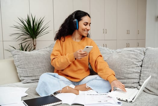Happy millennial arab student woman in wireless headphones writing learning notes, doing training course task, college homework, watching webinar, listening tutorials on laptop. Typing on the phone