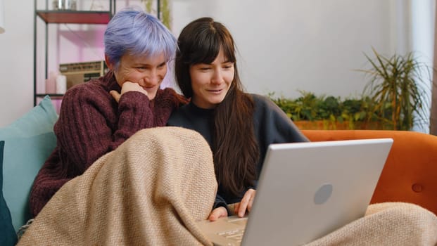 Two lesbian women couple or girls friends looking at laptop camera, making video webcam conference call with family, enjoying pleasant conversation, laughing, waving hello at home room. LGBT people