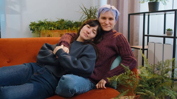 Two young lesbian women family couple at modern home living room. Loving same sex female gay girls hugging, relaxing together, lie on sofa. LGBT people. Romantic love relationship concept and emotion