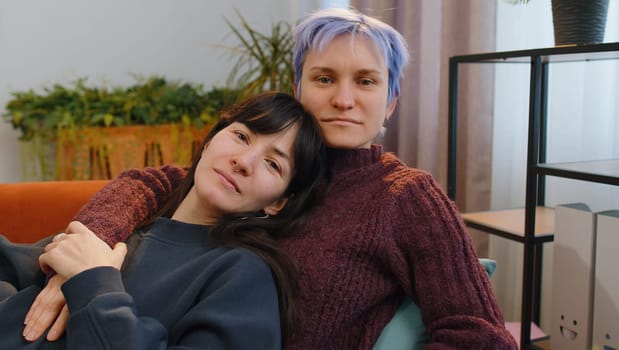 Two young lesbian women family couple at modern home living room. Loving same sex female gay girls hugging, relaxing together, lie on sofa. LGBT people. Romantic love relationship concept and emotion