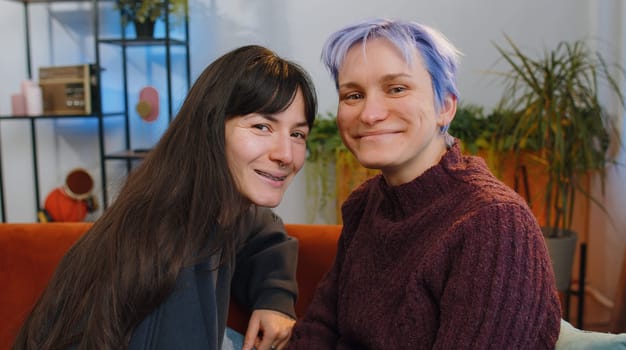 Two young lesbian women family couple looking at camera and smiling at home living room. Loving female gay girls hugging, relaxing together. LGBT people. Romantic love relationship concept and emotion