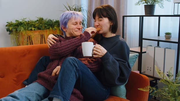Two lesbian women family couple, girl friends drinking coffee or tea, eating croissants and talking at home morning living room. LGBT same-sex people. Enjoying happy breakfast together lying on sofa