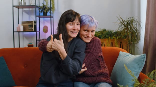 Like. Happy two young lesbian women family couple or girls friends looking approvingly at camera showing thumbs up, like sign something good positive feedback at home living room. LGBT people concept