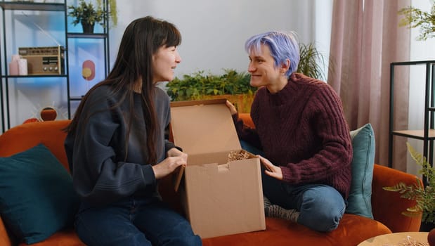 Happy two young lesbian women family couple unpacking delivery parcel at home living room. Smiling satisfied girls friends shoppers, online shop customers opening cardboard box receiving purchase gift