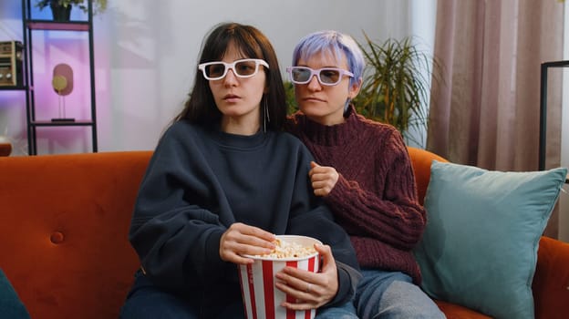 Excited two young lesbian women family couple or girls friends eating popcorn and watching interesting tv serial, sport game, film, online social media movie content at home living room. LGBT people