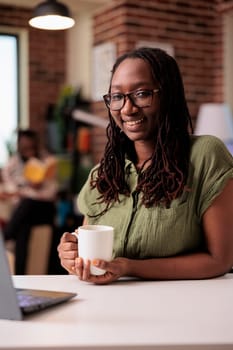 Portrait of smiling student with glasses relaxing having a coffee or tea while watching online series on laptop at home. Freelancer working remote taking a break while colleague is reading a book.