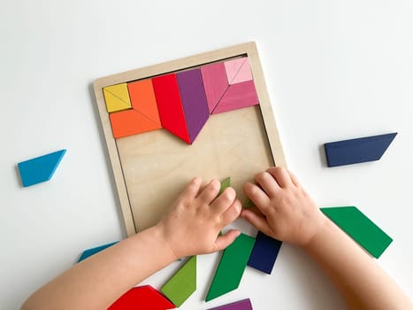childs hand collects multicolored wooden mosaic on white background. child solves a colorful tangram. square of colorful geometric shapes on white background.
