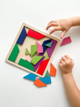 childs hand collects multicolored wooden mosaic on white background. child solves a colorful tangram. square of colorful geometric shapes on white background.