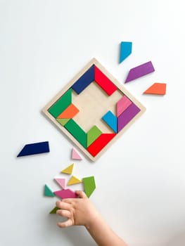 childs hand collects multicolored wooden mosaic on white background. child solves a colorful tangram. square of colorful geometric shapes on white background.