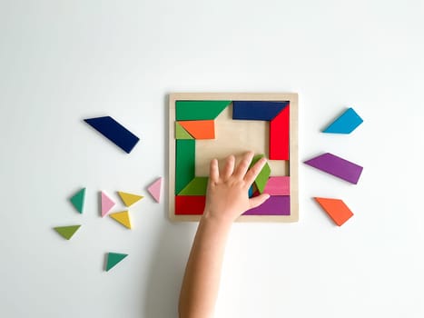 childs hand collects multicolored wooden mosaic on white background. child solves a colorful tangram. square of colorful geometric shapes on white background.